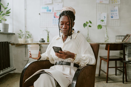 African American businesswoman drinking coffee and checking social network while resting in leather armchairの写真素材