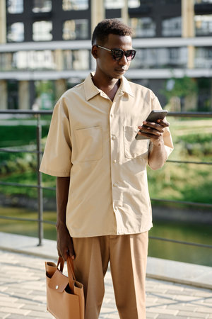 Vertical medium long shot of young African American male office worker wearing smart casual clothes standing outdoors looking at smartphone screenの写真素材