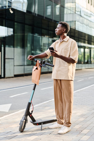 Vertical long shot of young African American man wearing smart casual outfit holding smartphone standing with his e-scooter outdoorsの写真素材