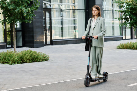 Wide shot of modern gen Z female office worker wearing fashionable formal outfit riding e-scooter, copy spaceの写真素材