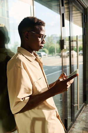 Vertical side view shot of young Black man wearing smart casual clothes spending break time outdoors surfing Internet on digital tabletの写真素材