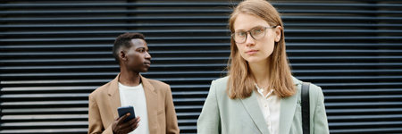 Header image of young Caucasian female manager holding smartphone and looking at camera, her Black male colleague standing relaxed in backgroundの写真素材