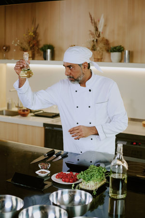 Indian male chef holding glass bottle with apple vinegar and looking at it while working in professional kitchenの写真素材