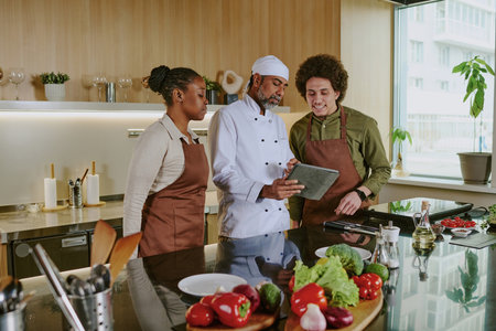 Professional chef holding touchpad with new menu and discussing it with his assistants while they standing in center of kitchenの写真素材