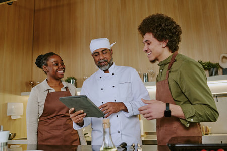 Two young cooks and mature highly skilled chef with touchpad in hand discussing new menu in modern kitchenの写真素材