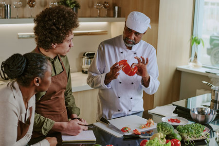 High angle shot of professional Indian chef holding cut red bell pepper and giving interns advice about vegetable cuttingの写真素材