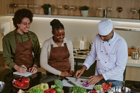 High angle shot of chef in white uniform cutting red onion while his biracial interns writing down his actionsの写真素材
