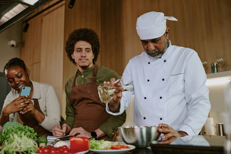 Low angle shot of Indian chef adding vinegar while interns recording video and watching him in modern kitchenの写真素材