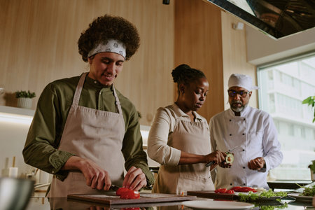 Low angle shot of curly haired cook cutting tomato while his African American colleague peeling avocado, Indian chef in white uniform watching process and giving advicesの写真素材