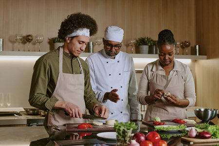 Highly skilled chef giving advices about cooking process, biracial interns preparing ingredients and listening to their masterの写真素材