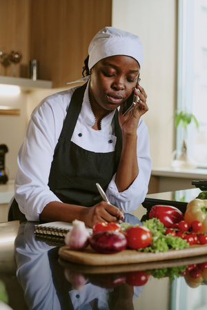 Vertical shot of African American cook speaking on phone with client and writing down his desires of upcoming celebration menu in this restaurantの写真素材