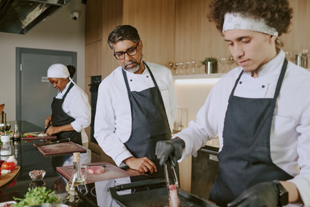 Mature Indian chef controlling his young cook while he flipping steak, female cook cutting vegetables in backgroundの写真素材