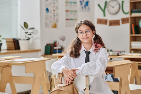 Medium portrait of modern teen girl wearing uniform and eyeglasses sitting on chair in classroom and looking at cameraの写真素材
