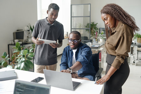 Young African American teamlead with disability pointing to laptops screen while his coworker looking at itの写真素材