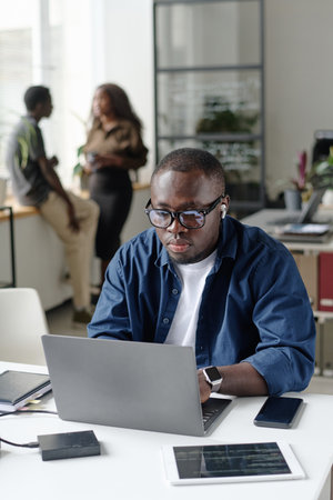 Vertical shot of African American man sitting at work desk and typing on keyboard while listening to musicの写真素材