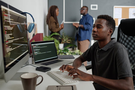 Young African American app developer sitting at white work desk with computer, coffee cup and laptop, he writing codes while his colleagues chattingの写真素材
