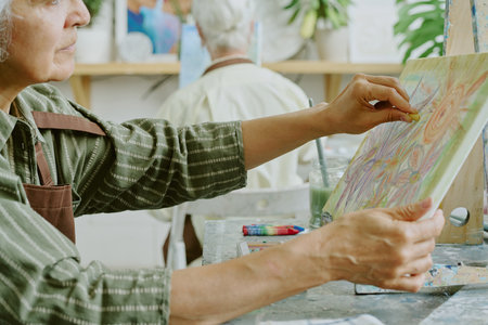 Side view shot of senior woman holding canvas with painted flowers while adding details with yellow pastel chalkの写真素材