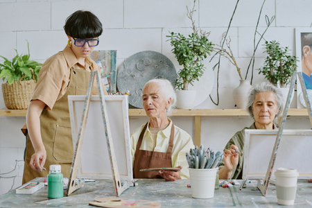 Young adult female teacher with bob haircut checking paintings on canvas of senior studentsの写真素材