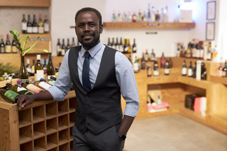 Medium close up of African American sommelier leaning on wooden rack with bottle of wineの写真素材