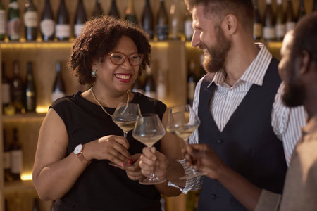 Mixed race beautiful woman smiling and clinking glasses full of white wine with her colleagues during degustation in wine cellarの写真素材
