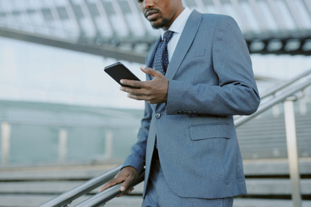 Professional African American man in suit holding smartphone while standing on staircase in modern building. Background includes architectural details of the structureの写真素材