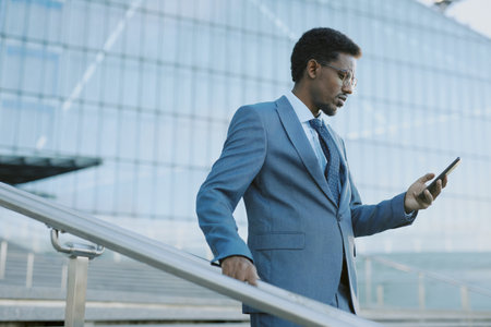 Businessman in blue suit checking smartphone while standing on stairs outside modern glass building during daytime. Background includes architectural structure with metal railingsの写真素材