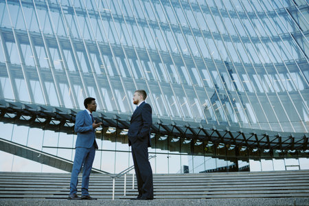 Two businessmen standing and conversing in front of a contemporary glass building with a unique architectural design in the backgroundの写真素材
