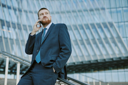 Caucasian businessman wearing suit talking on phone while standing on stairs outside modern glass office building with serious expressionの写真素材