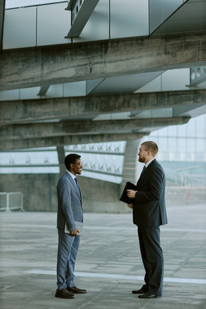 Two businessmen are standing in conversation in modern office building. Both wearing suits, they are engaged in professional discussion in contemporary architectural surroundingsの写真素材