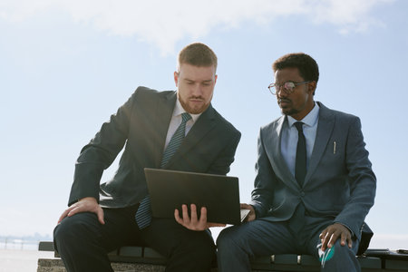 Two businessmen having collaboration while using laptop outdoors during sunny day, sharing discussion. Focus is on interaction between colleagues in professional attireの写真素材