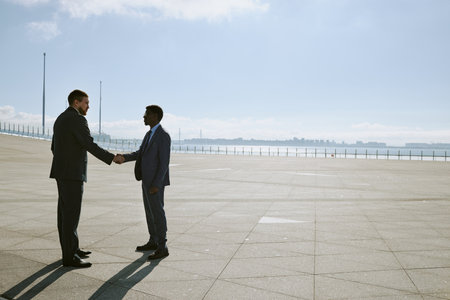 Two business professionals shaking hands standing in a spacious outdoor area surrounded by large cityscape on horizon. Background features a vast, open space under clear blue skyの写真素材