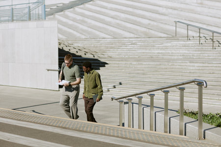 Two men walking down concrete staircase outdoors, one handing over a document. Bright sunlight creating shadows adds depth to the sceneの写真素材