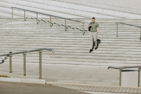 Businessman in green shirt and trousers walking down wide outdoor stairs carrying bag in hand. Clear day with sunlight illuminating stairs and railings in backgroundの写真素材