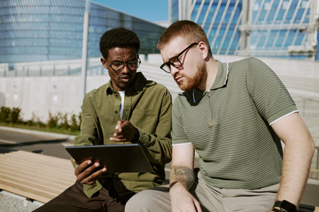 Two young men sitting outdoors, discussing project on a tablet. Background includes modern architectural elements and clear sky, suggesting a productive, open environmentの写真素材