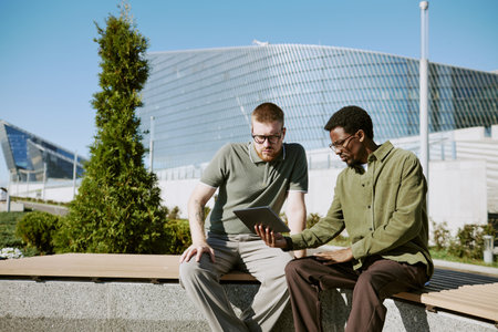 Two men discussing project on tablet while sitting on bench in modern urban area with glass architecture in backgroundの写真素材