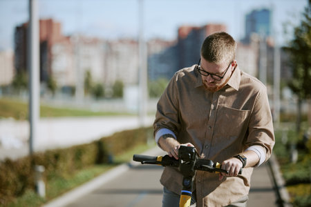 Man riding electric scooter on pathway within urban area, focusing on handlebar while background showcases blurred city buildings on sunny dayの写真素材
