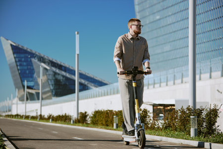 Man riding an electric scooter on pavement near modern architectural structures under clear blue sky, holding handlebars firmly, and staring into the distanceの写真素材