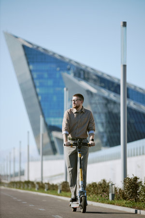 Man riding electric scooter near a modern angular building exhibiting contemporary architectural style on a clear dayの写真素材