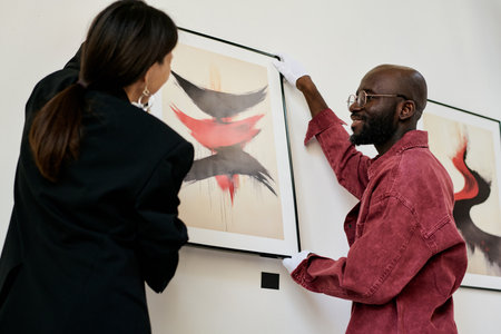 Man wearing white gloves and red shirt hanging framed art while woman observing. Abstract art pieces feature dynamic brush strokes with bold color contrasts framing gallerys wallの写真素材