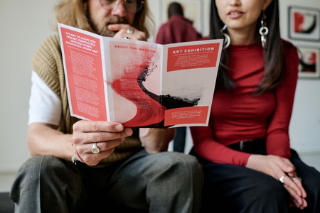 Two people sitting closely together discussing details of an art exhibition pamphlet, with additional background figures visible. Casual yet focused atmosphere shared by individualsの写真素材