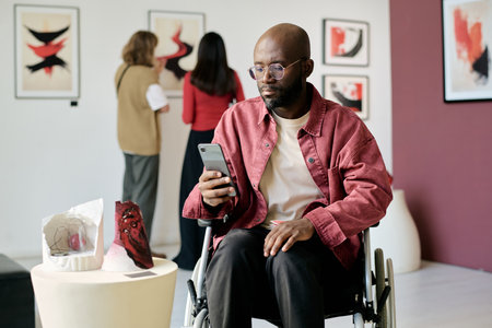 Man in wheelchair looking at smartphone while visiting an art exhibit, with artwork and people visible in background, creating a thoughtful and contemplative atmosphereの写真素材