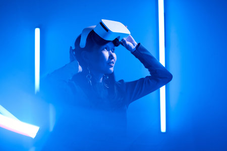 Woman experiencing virtual reality, adjusting her headset in a dimly lit room surrounded by blue...