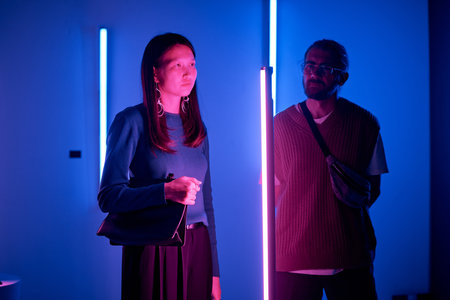 Portrait of woman illuminated by colorful neon lights, standing in a dimly lit room with a man in the background looking at her, creating a moody atmosphereの写真素材