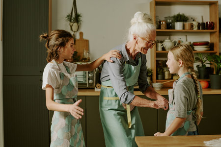 Elderly woman teaching two young girls how to bake while smiling and wearing aprons in cozy kitchen setting with various cooking utensils and ingredients aroundの写真素材