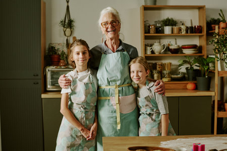Senior woman baking with two young girls, wearing aprons and smiling together in kitchen, standing near wooden table with baking suppliesの写真素材