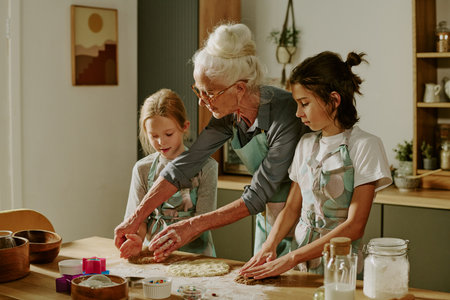 Senior woman guiding two young girls in kitchen while preparing dough together, focused on baking cookies and enjoying the moment without distractionsの写真素材