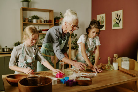 Senior woman teaching two young girls to bake cookies in cozy kitchen with wooden table and various baking utensils. Children focused on cutting out shapes from dough with colorful moldsの写真素材