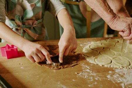 Child and adult working together on dough, cutting shapes using cookie cutter on wooden surface. Hands covered with flour and raw dough cut into various formsの写真素材