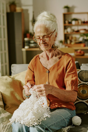 Elderly woman sitting on sofa crocheting in cozy living room filled with plants and shelves in backgroundの写真素材