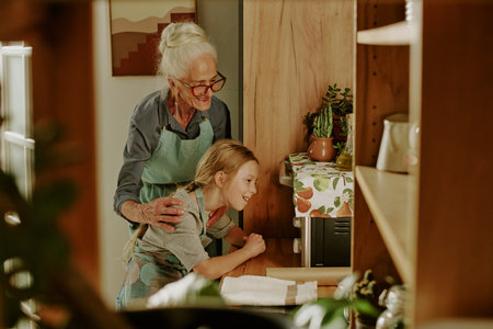 Senior woman and young girl cooking in warmly lit kitchen, enjoying quality time together. They are wearing aprons and sharing a moment of joy while preparing foodの写真素材